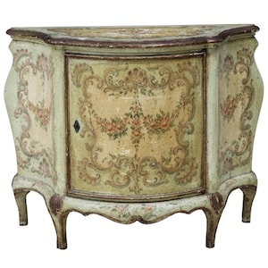 May include: A small, antique cabinet with a curved front and ornate floral designs. The cabinet is painted in shades of green and cream, with a distressed finish. The door features a central floral motif, and the legs are curved.