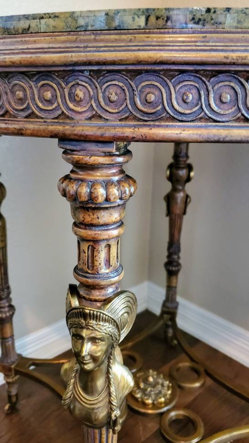 May include: Ornate antique table with a marble top and detailed carvings. The table features a bronze bust of a woman and decorative brass accents. The legs are intricately designed with classical motifs, showcasing a blend of materials and craftsmanship.
