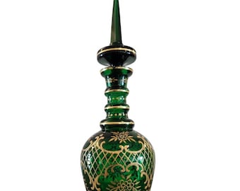 May include: A tall, green glass decanter with a gold-painted design. The decanter has a narrow neck and a pointed stopper.