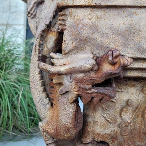 May include: A rusty metal dragon sculpture with intricate details. The dragon is holding a rectangular planter with a weathered surface. The sculpture is a garden ornament.