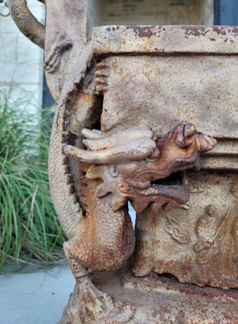 May include: A rusty metal dragon sculpture with intricate details. The dragon is holding a rectangular planter with a weathered surface. The sculpture is a garden ornament.