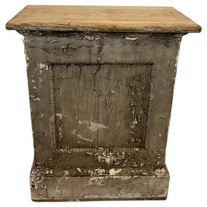 May include: A distressed gray wooden pedestal with a square top. The pedestal has a single door with a raised frame and a simple design. The paint is chipped and faded, giving it a vintage look.