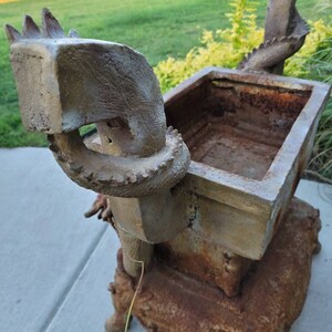 May include: A rusty metal planter shaped like a dragon with a square planter box on top. The planter is decorated with a scaly texture and has a weathered, aged look.