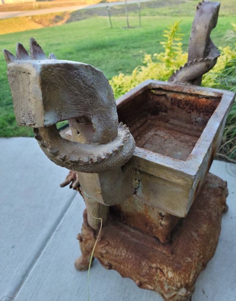 May include: A rusty metal planter shaped like a dragon with a square planter box on top. The planter is decorated with a scaly texture and has a weathered, aged look.
