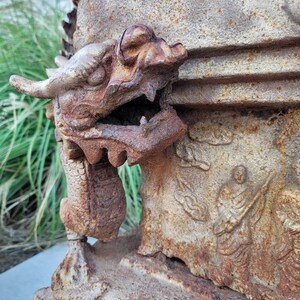 May include: A rusty metal dragon sculpture with a detailed head and open mouth. The sculpture is attached to a larger, weathered stone structure with carvings.