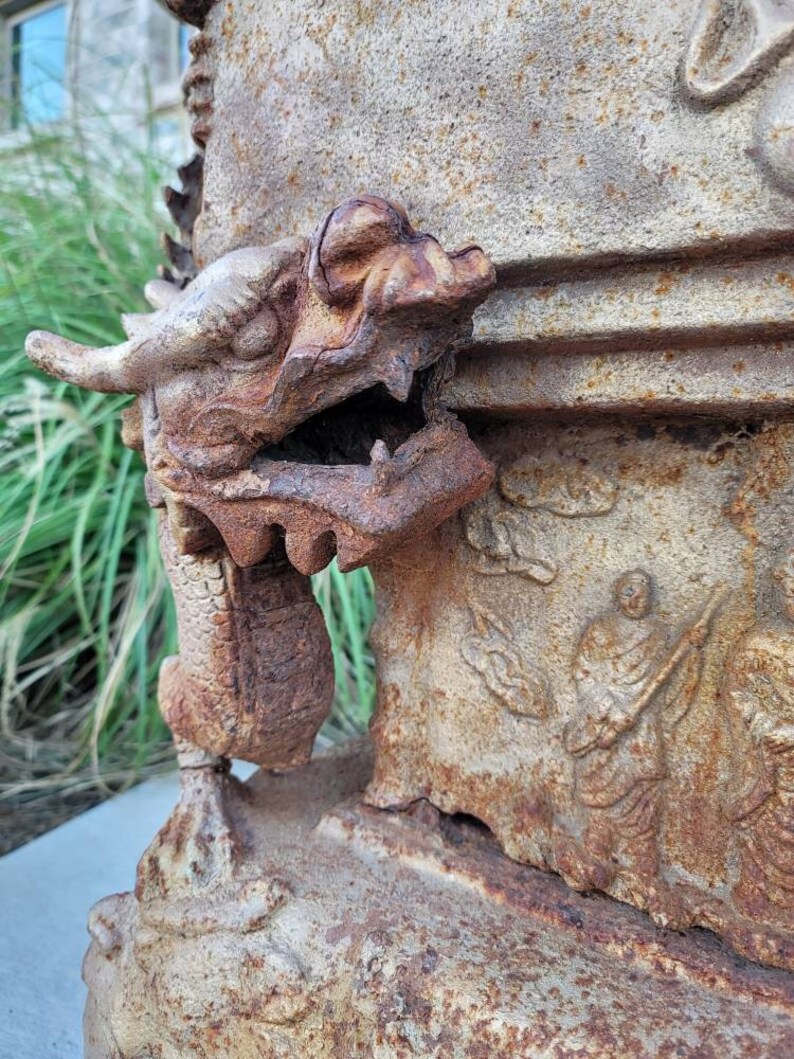 May include: A rusty metal dragon sculpture with a detailed head and open mouth. The sculpture is attached to a larger, weathered stone structure with carvings.