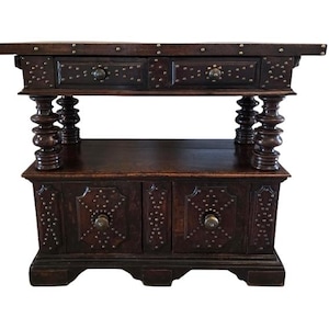 May include: Dark brown wooden cabinet with two drawers and two doors. The cabinet is decorated with small metal studs and ornate hardware. The legs are turned and the cabinet has a shelf.