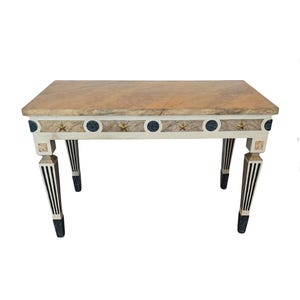 May include: A rectangular table with a light brown marble top. The table's apron is white with blue circles, gold stars, and marble accents. The legs are black and white striped with black bases.