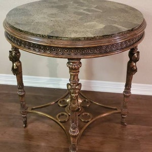 May include: A round antique side table with a marble-like top and ornate gold-colored legs. The table has a decorative edge and detailed carvings, showcasing a classic design. The table's height is approximately 24 inches.