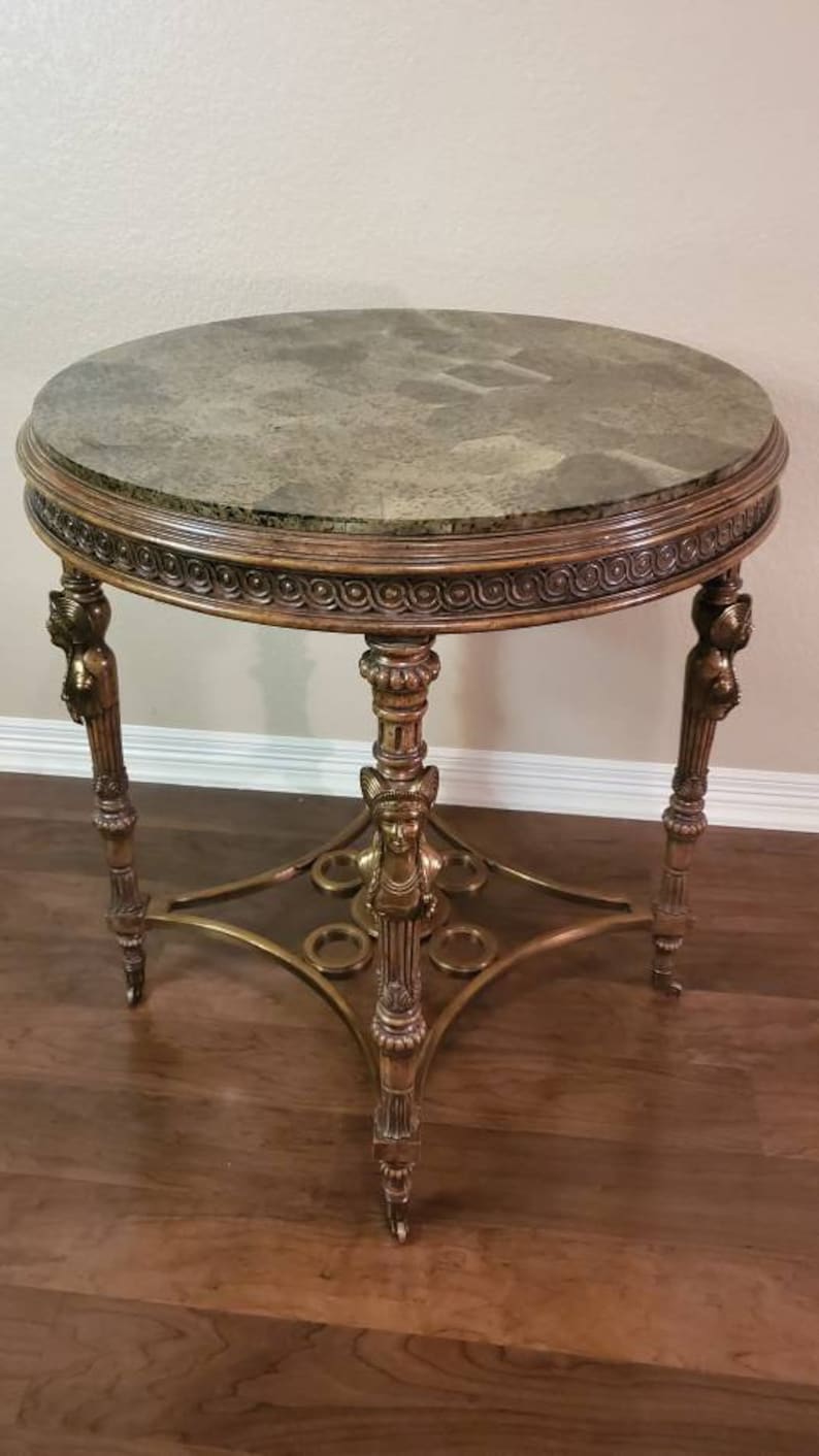 May include: A round antique side table with a marble-like top and ornate gold-colored legs. The table has a decorative edge and detailed carvings, showcasing a classic design. The table's height is approximately 24 inches.