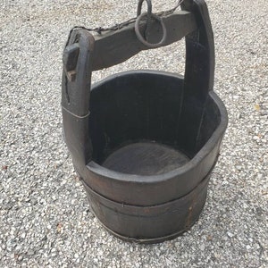 Puede incluir: Un cubo de madera marr&oacute;n oscuro con un asa de metal y una cuerda atada al asa. El cubo est&aacute; sentado sobre una superficie de grava.