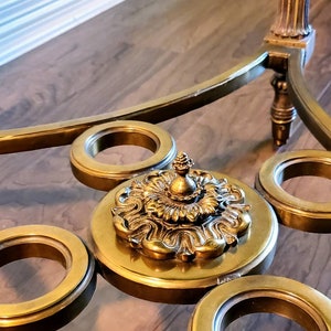 May include: A close-up of a gold-toned, ornate coffee table. The table features a glass top with decorative gold rings and a central, detailed medallion. The legs and frame are also gold, creating a cohesive, elegant design.