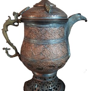 May include: An antique copper teapot with intricate floral designs and a brass handle. The teapot has a lid with a brass knob and sits on a decorative stand with an openwork design.