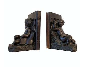 Puede incluir: Un par de sujetalibros con una escultura de bronce de un querubín. Cada querubín está sentado en un libro y sostiene un libro en sus manos. Los sujetalibros son de color marrón oscuro.