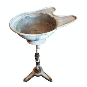 May include: An antique, white metal shampoo bowl on a tripod stand. The bowl has a unique shape with a contoured neck rest. The stand is adjustable and has a three-legged base. The metal shows signs of age and wear.