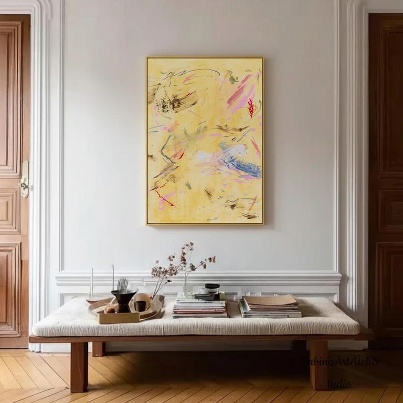 May include: Abstract art piece with a gold frame. The artwork features a yellow background with colorful brushstrokes in pink, red, blue, and black. The painting hangs above a wooden bench with books and decorative items.