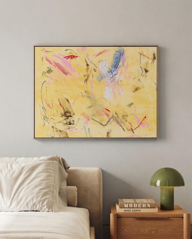 May include: Abstract art piece with a yellow background and colorful brushstrokes in pink, blue, and red. The artwork is framed in a thin, dark brown frame and hangs on a light gray wall. A bed with white linens and a nightstand with a green lamp are in the foreground.