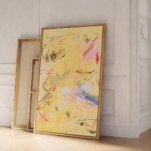 May include: Abstract art piece with a yellow background and colorful brushstrokes in pink, blue, and red. The artwork is framed in a natural wood frame and leans against a white wall. Additional unframed canvases are visible.