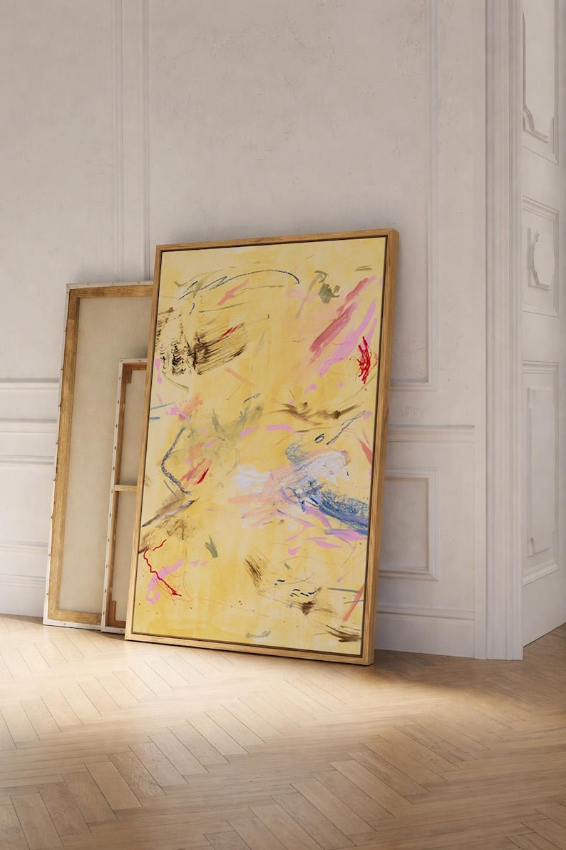 May include: Abstract art piece with a yellow background and colorful brushstrokes in pink, blue, and red. The artwork is framed in a natural wood frame and leans against a white wall. Additional unframed canvases are visible.