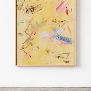May include: Abstract painting on canvas with a light yellow background. The artwork features brushstrokes in shades of pink, blue, red, and brown. The painting is framed in a light wood frame and is displayed against a white wall.