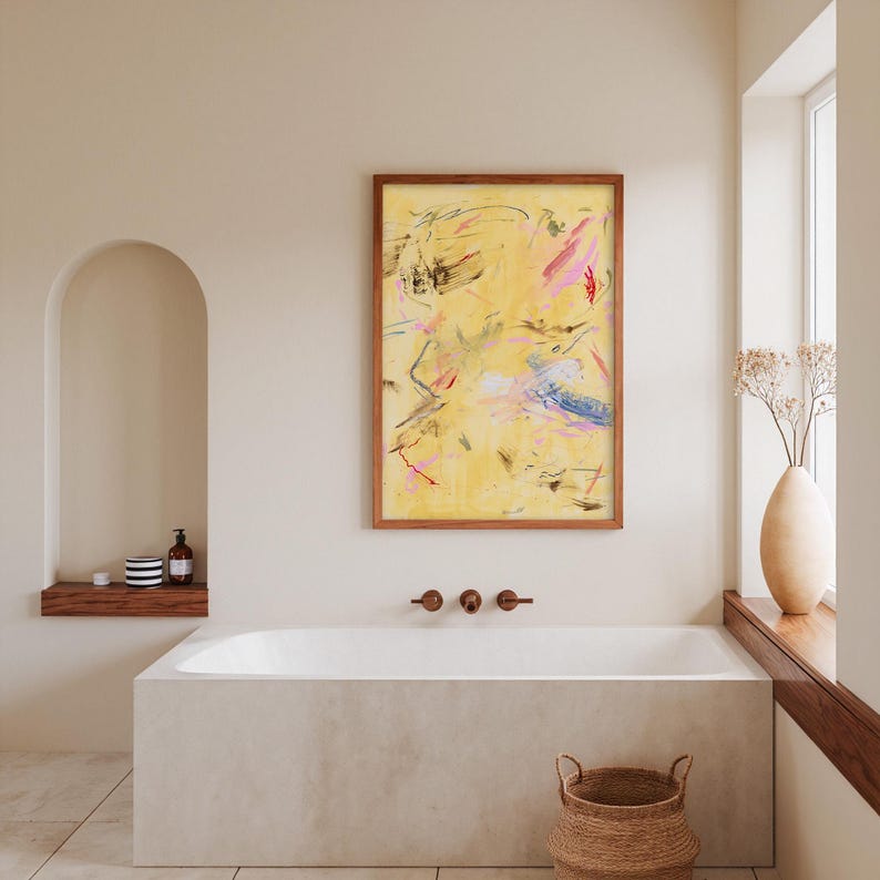 May include: Abstract art piece in a wooden frame. The artwork features a yellow background with brushstrokes of pink, blue, and black. The painting hangs above a white bathtub in a bathroom setting. A woven basket sits on the floor.