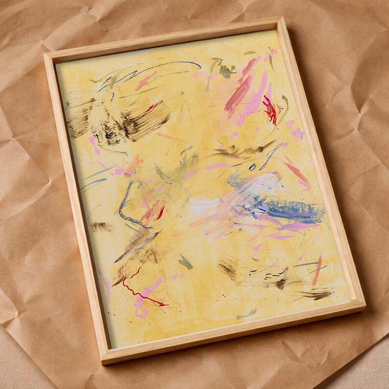 May include: Abstract art print in a wooden frame. The artwork features a yellow background with colorful brushstrokes in pink, blue, red, and brown. The framed art is displayed on a brown paper surface.