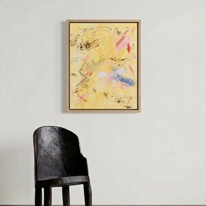 May include: Abstract art piece in a wooden frame. The artwork features a yellow background with expressive brushstrokes in pink, black, and blue. A dark wooden chair is in the foreground.