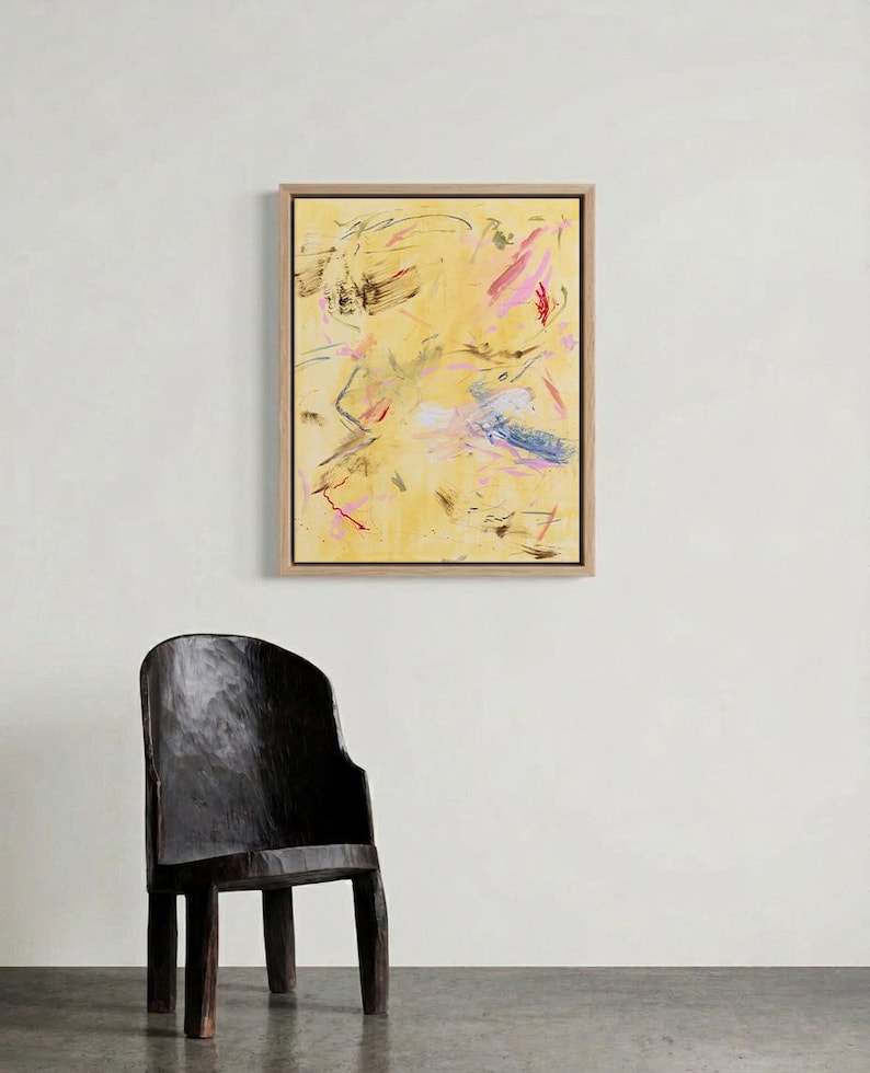 May include: Abstract art piece in a wooden frame. The artwork features a yellow background with expressive brushstrokes in pink, black, and blue. A dark wooden chair is in the foreground.