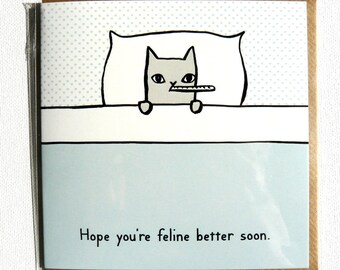 May include: A greeting card featuring a cartoon cat in bed with a thermometer in its mouth. The cat is gray with black outlines, resting on a white pillow and under a light blue blanket. The text "Hope you're feline better soon" is at the bottom.