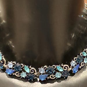 Könnte beinhalten: Eine silberfarbene Halskette mit floralem Design, die blaue und türkisfarbene Edelsteine aufweist. Die Halskette wird auf einer schwarzen Schaufensterpuppe präsentiert, die die komplizierten Details und Farbvariationen der Steine hervorhebt.