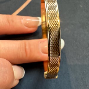 May include: A gold-coloured bangle bracelet with a textured, patterned design. The bracelet has a rectangular clasp and a repeating wave-like pattern along the top edge. The bracelet is shown against a dark background.