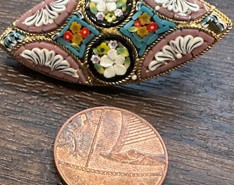 May include: An oval mosaic brooch with a gold-tone frame. The brooch features a pink background with white and teal accents, and circular and diamond-shaped floral designs. A penny is next to the brooch.