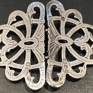 May include: Ornate silver-toned clasp with a floral design. The clasp is composed of two symmetrical halves, each featuring a central oval and intricate, swirling patterns. The metal has a slightly aged appearance.