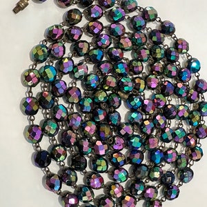 May include: A long strand of iridescent glass beads in shades of purple, blue, and green. The beads are faceted and have a metallic sheen.
