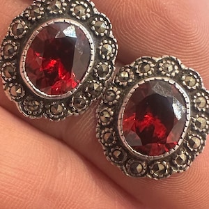 May include: A pair of silver earrings with red gemstone centres surrounded by a border of small, black marcasite stones.
