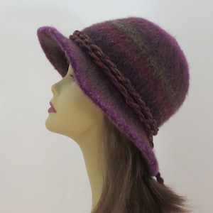 May include: A purple, brown, and green wool felt hat with a braided trim. The hat has a wide brim and a rounded crown.