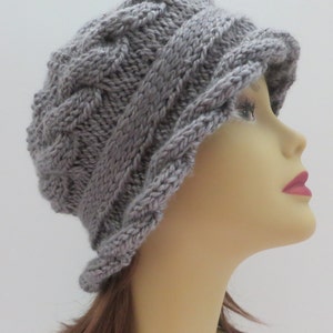Puede incluir: Un gorro de punto gris con un patrón de trenza. El gorro tiene un ala ancha y está doblado hacia abajo en un lado.