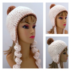 May include: A white knitted hat with brown trim and a brown pom-pom on top. The hat has ear flaps that are tied with a knot at the end.