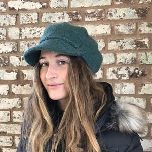 Puede incluir: Una mujer con una gorra de estilo newsboy azul verdoso, una chaqueta negra y un ribete de piel sintética. La gorra tiene un detalle trenzado alrededor del borde.