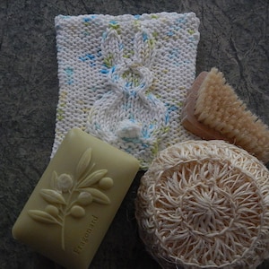 May include: A white knitted dishcloth with a bunny design, a bar of soap with an olive branch design, a natural loofah, and a wooden brush with natural bristles.