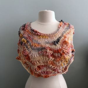 May include: A crocheted shawl in shades of orange, pink, and grey. The shawl has a delicate, lacy pattern and is displayed on a white mannequin.