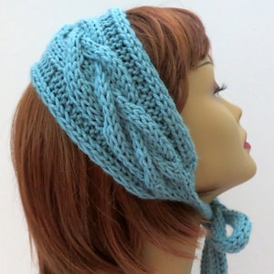 Puede incluir: Una diadema de punto azul claro con un patrón de punto trenzado y una larga corbata. La diadema se lleva en un maniquí con el pelo rojo.