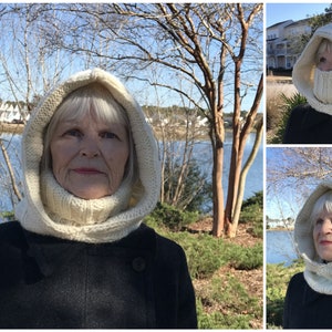 Puede incluir: Una bufanda de punto blanca con capucha y cuello de tortuga. La bufanda la lleva puesta una persona de pie frente a un lago y árboles.