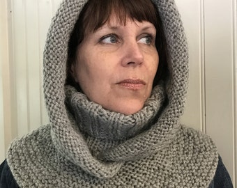 May include: A gray knitted cowl neck with a hood. The cowl is folded down and the hood is up. The cowl is worn by a person with brown hair.
