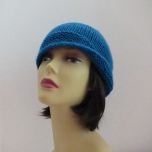 May include: A teal blue knitted hat with a turned-up brim. The hat is made from a soft, textured yarn and is designed to fit snugly. The hat is a solid colour and appears to be handmade.