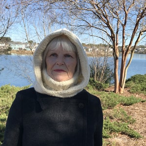May include: A woman wearing a white knitted hooded scarf and a black coat. She is standing in front of a body of water with trees in the background.