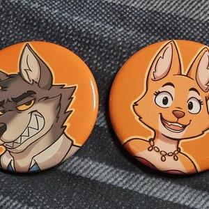 Könnte beinhalten: Zwei runde, orangefarbene Buttons mit Cartoon-Zeichnungen von anthropomorphen Wolfs- und Fuchscharakteren. Der Wolf hat einen verschmitzten Gesichtsausdruck und der Fuchs lächelt. Die Buttons sind vor einem Jeans-Hintergrund platziert.