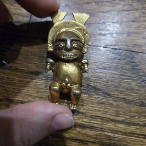 May include: A small gold figurine of a person with a stylized face and arms outstretched. The figurine is sitting on a wooden surface.