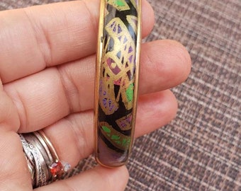 May include: A gold bangle bracelet with a black background and colorful abstract design. The design features gold, green, purple, and pink accents.