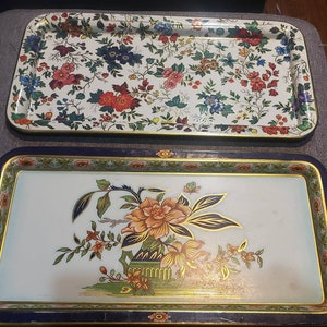 May include: Two vintage metal serving trays. The top tray has a white background with a floral pattern in shades of pink, red, green, and blue. The bottom tray has a white background with a gold floral design on a blue border.